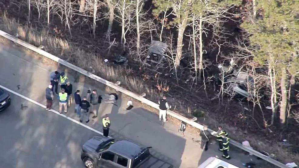 Aerial view of a falmouth car accident scene showing emergency responders and bystanders near a wooded roadside. Two wrecked vehicles are visible among the trees beyond a guardrail, indicating a high-speed off-road accident.
